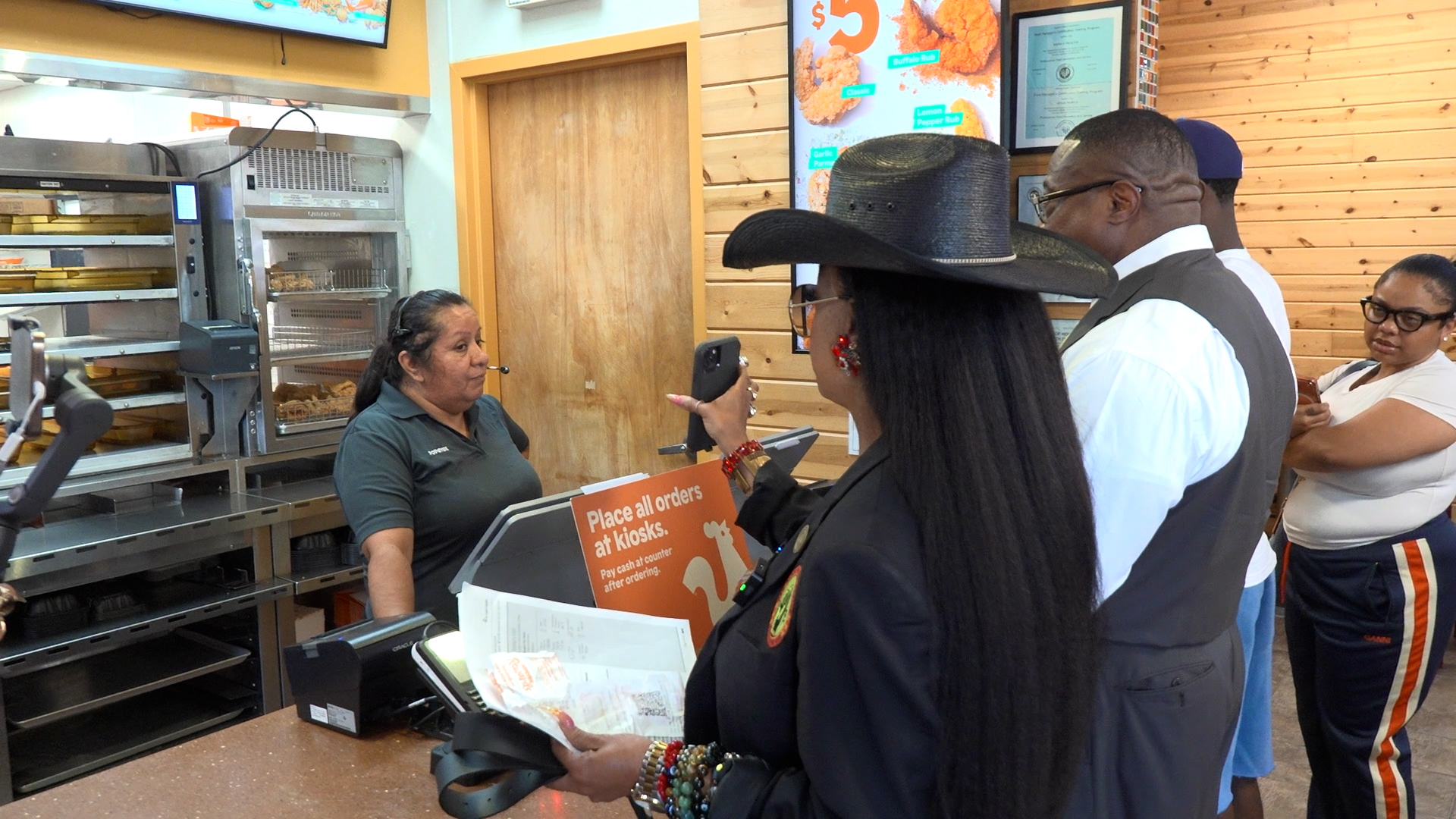 Group of customers at a fast-food counter, one woman in a black cowboy hat using a smartphone near a 'Place all orders at kiosks' sign.