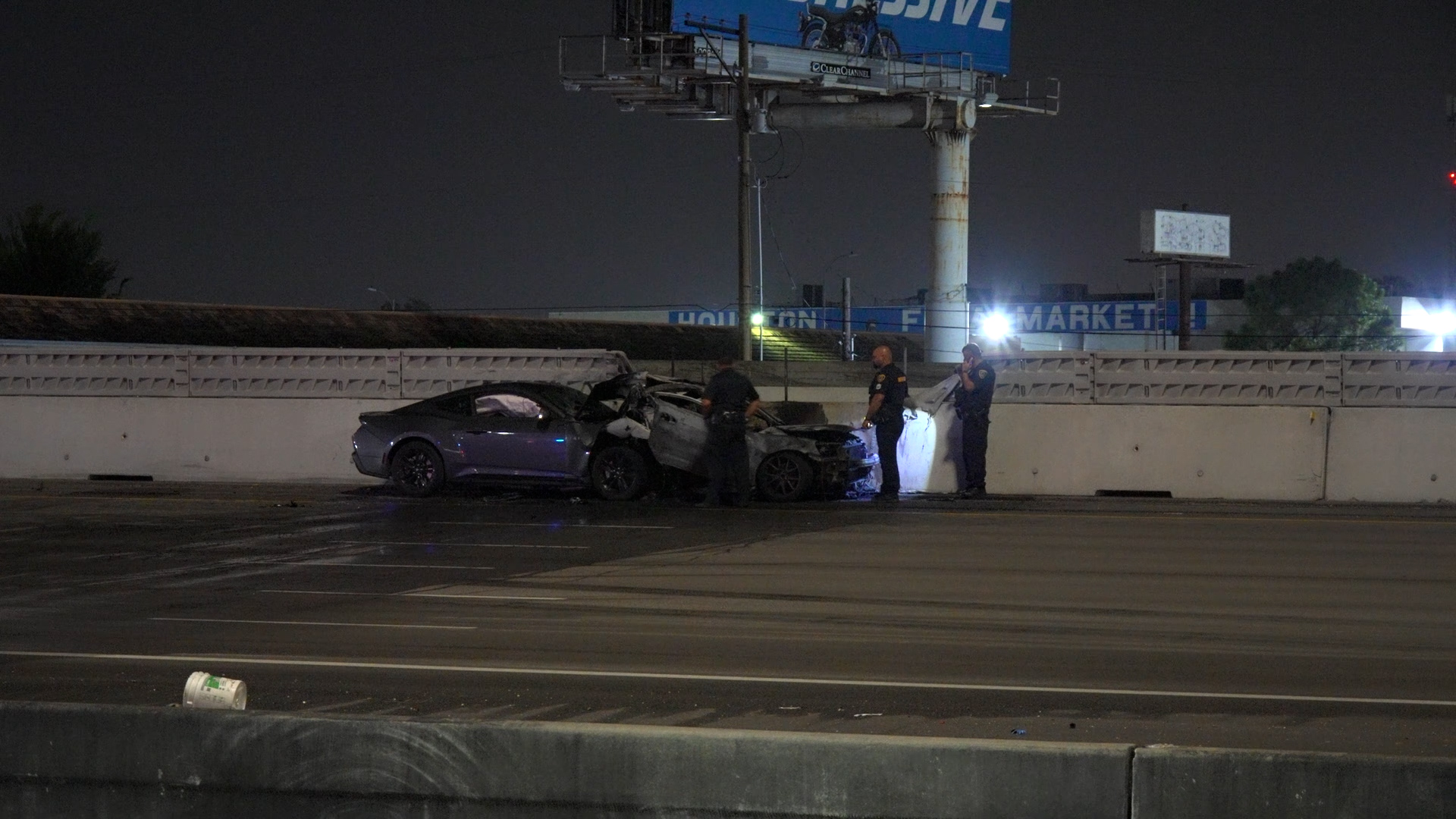 Nighttime highway crash scene with two damaged cars against a concrete barrier and three police officers examining the wreckage under overhead streetlights.