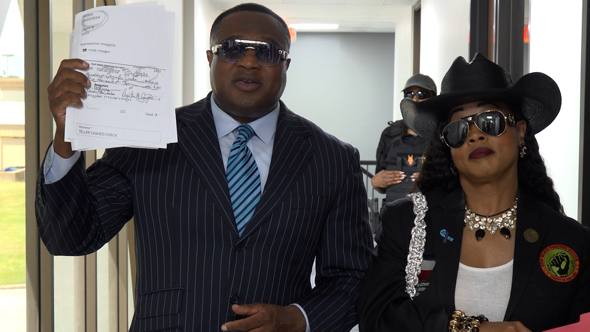 Man in a pinstripe suit wearing sunglasses holds a stack of papers as he and a woman in a black hat and big sunglasses walk through a doorway behind a security guard.