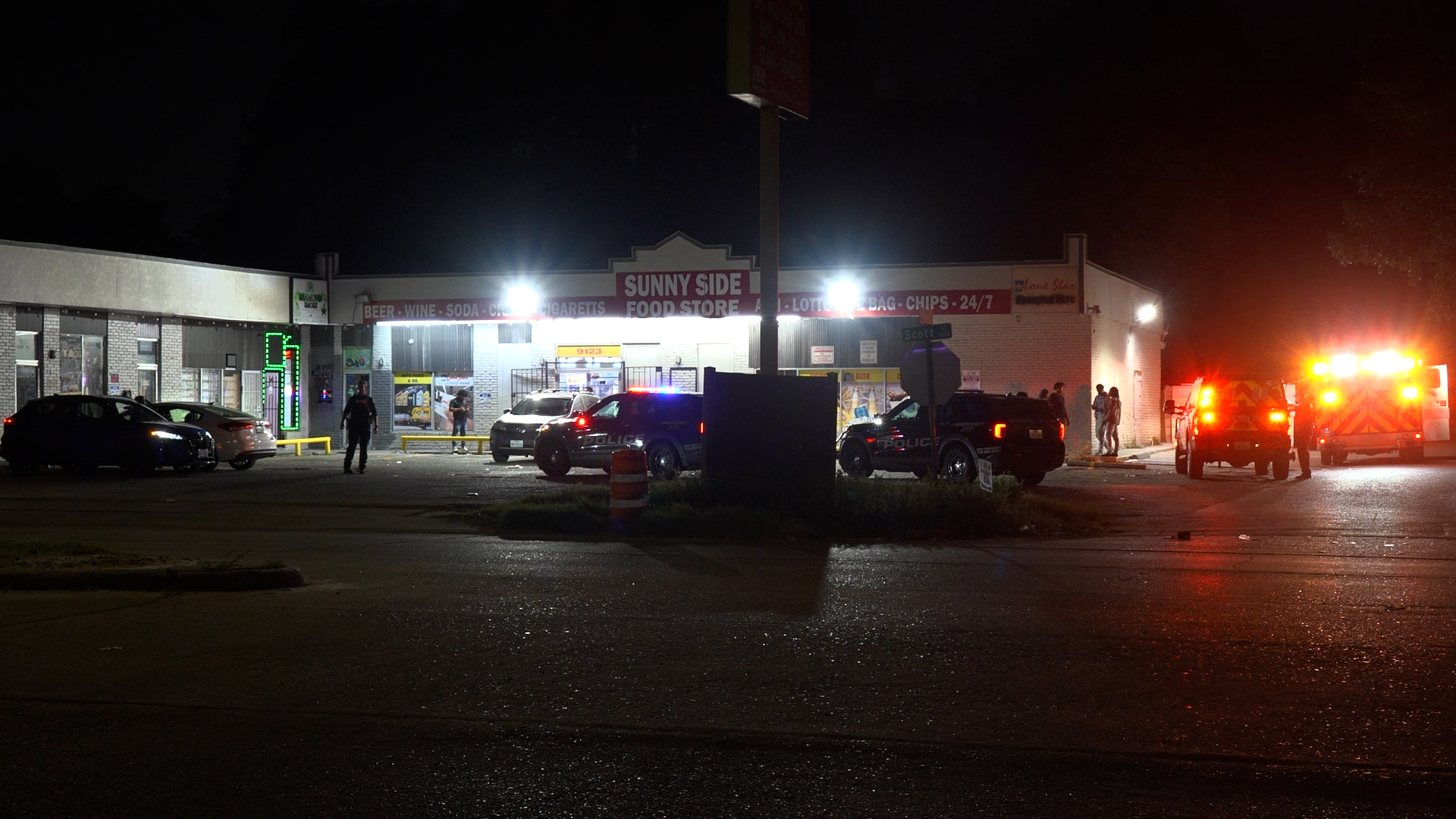 Night exterior of Sunny Side Food Store with police cars and an ambulance, flashing lights, and several bystanders outside the entrance at night.
