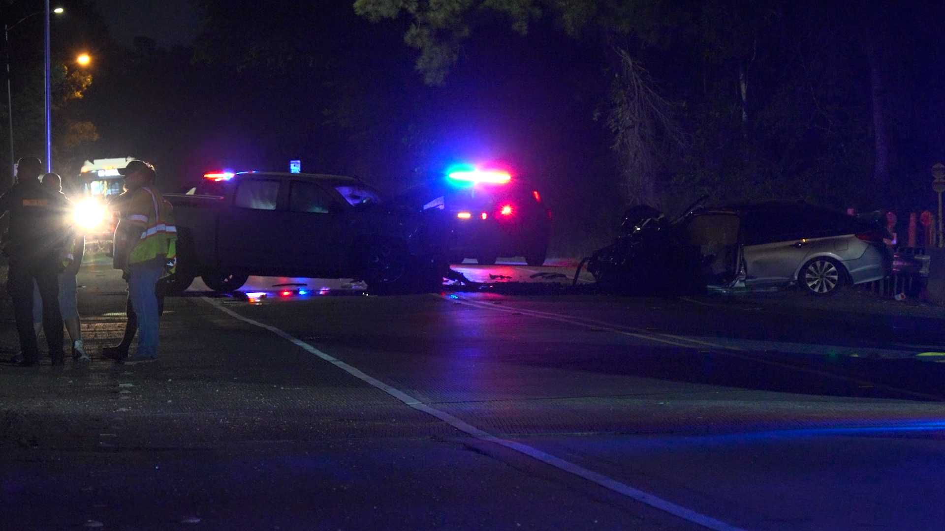 Nighttime traffic accident scene with flashing red and blue police lights, damaged sedan, and responders on a dark street edge.
