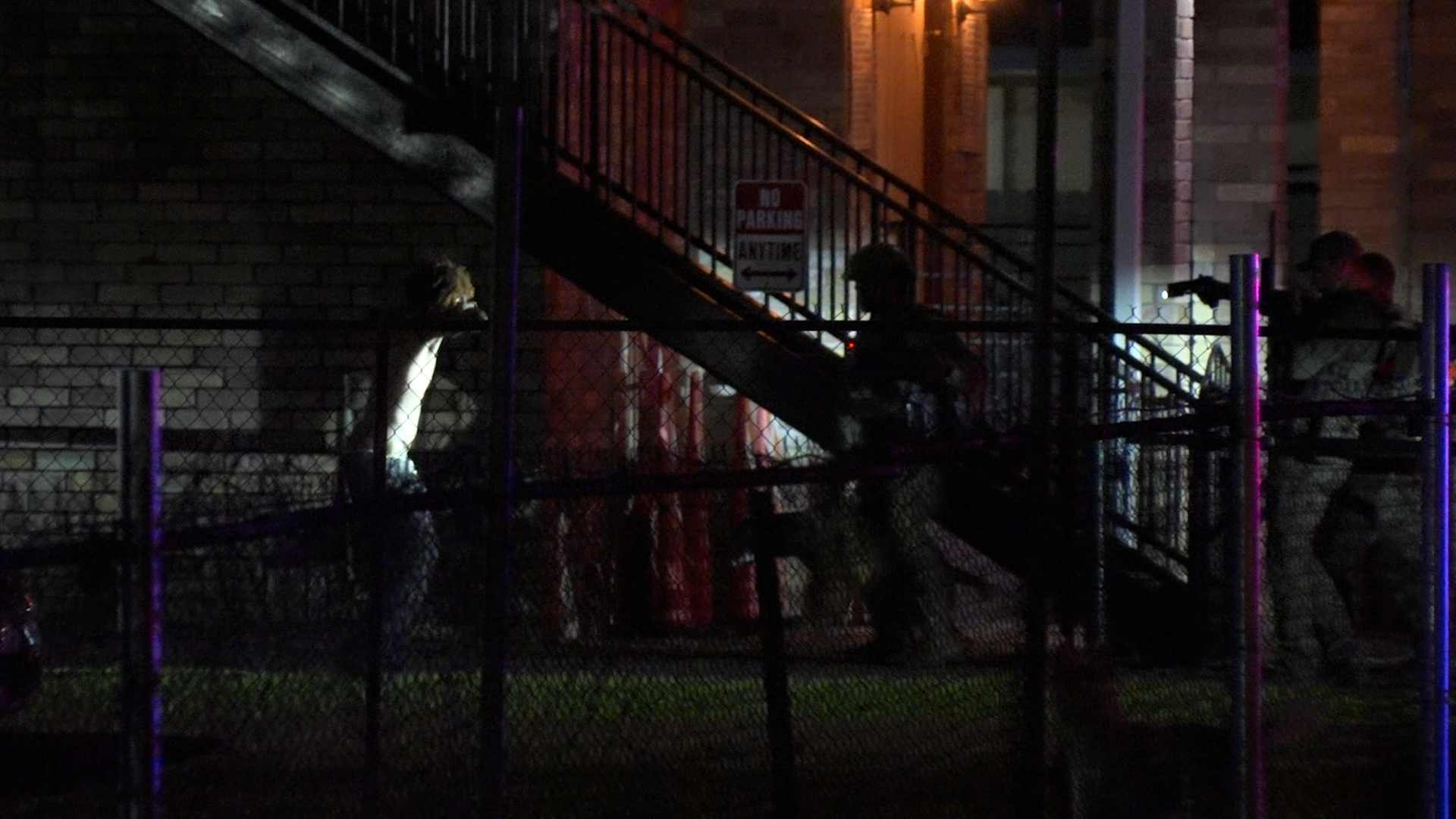 Nighttime urban scene with a metal staircase behind a chain-link fence and a silhouette of a person nearby, with someone holding a camera on the right.