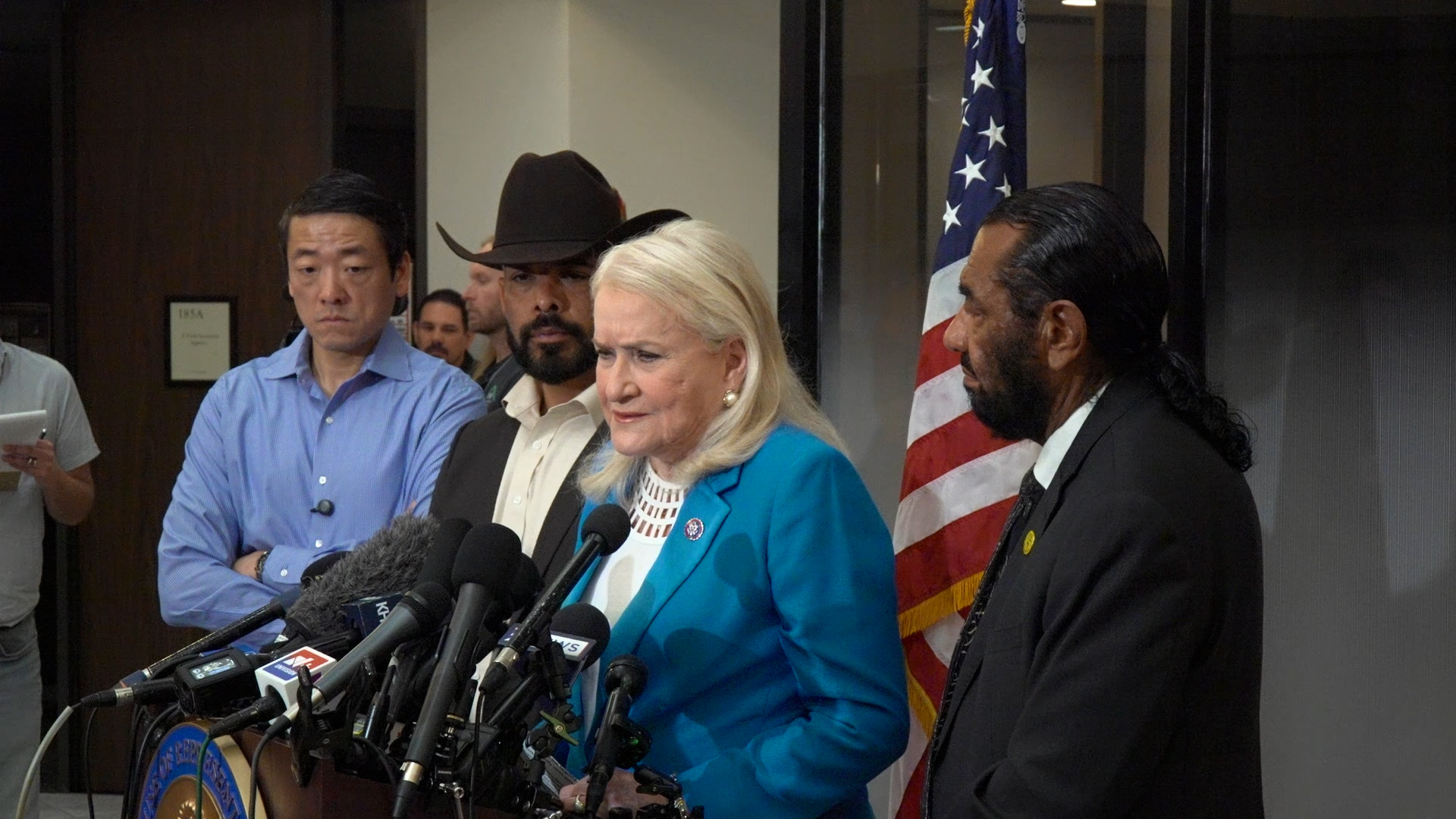 Speaker in a turquoise blazer addresses the media at a press conference, microphones in front and the U.S. flag behind her.