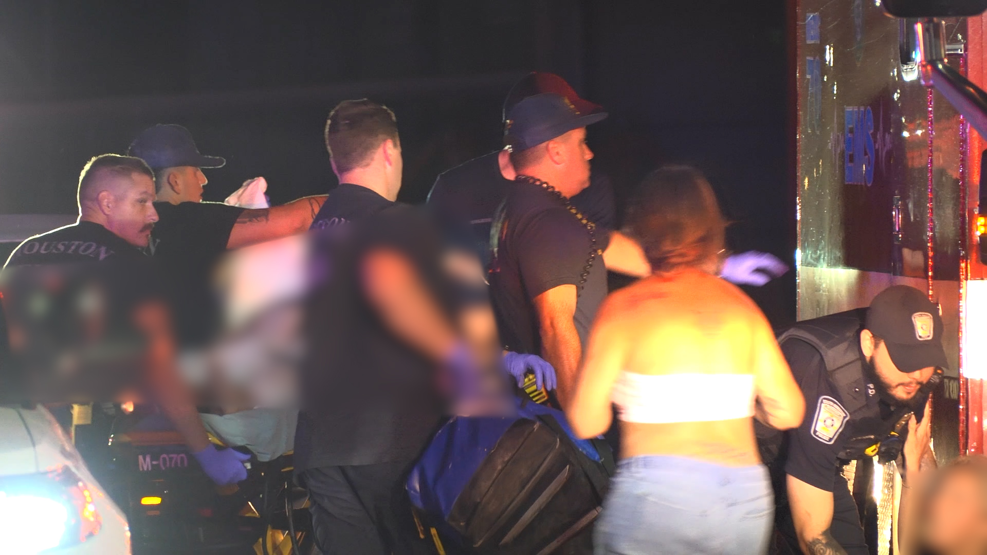 Group of emergency responders and bystanders at night beside a large reflective vehicle, some wearing gloves and utility belts, with a shirtless person in the foreground.