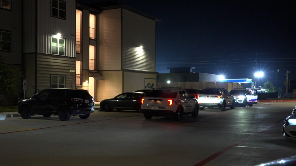 Nighttime parking lot in front of a modern apartment building with several SUVs parked along the curb; exterior lights are on.