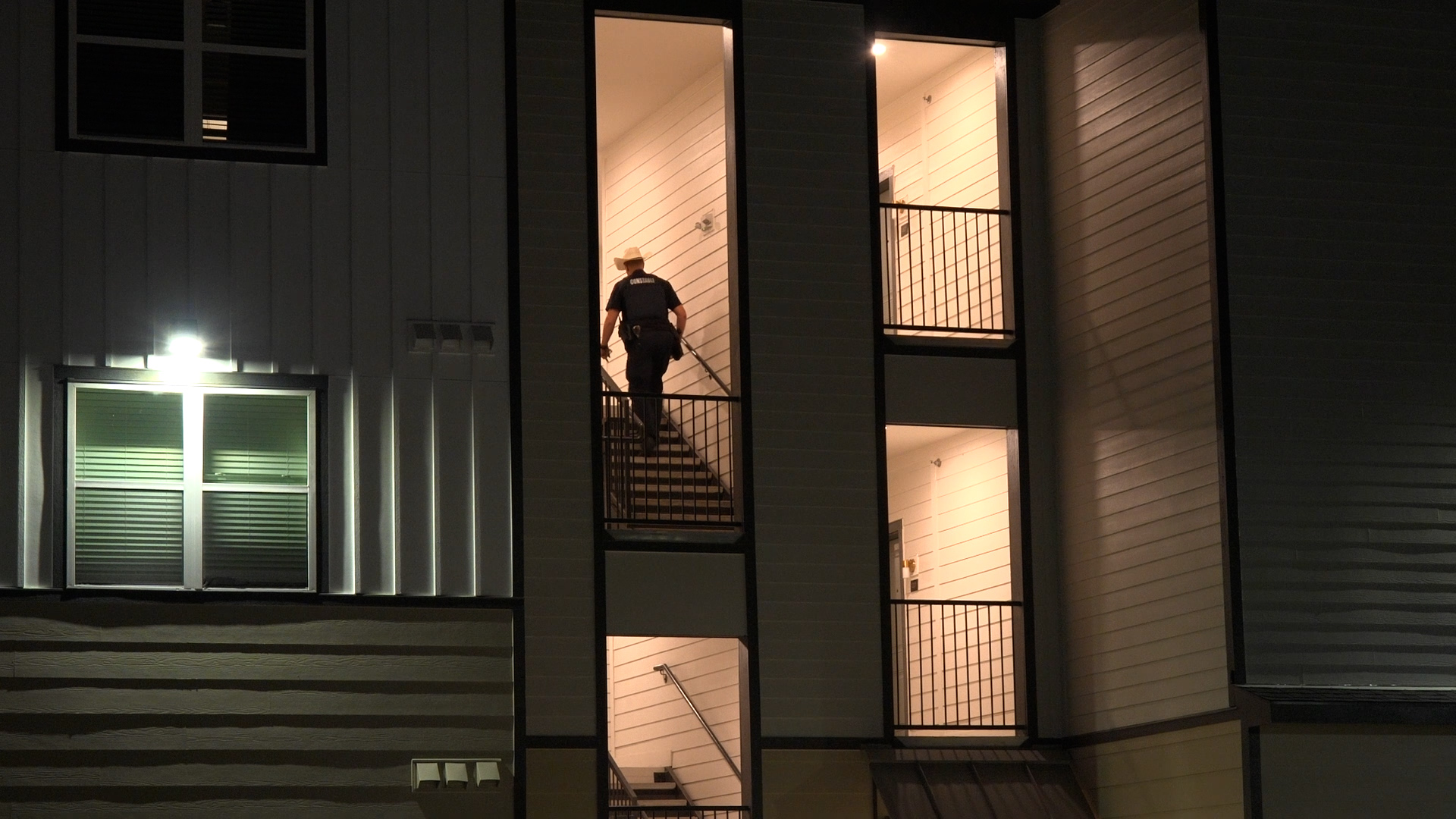 Night view of a multi-story building with a person in a dark uniform standing on a lit interior stairwell visible through tall windows at night.