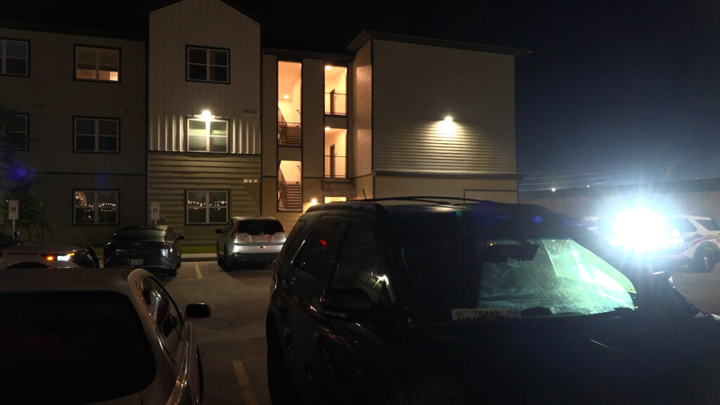 Nighttime view of a modern multi-story apartment building with illuminated stairwell windows, and several parked cars in a dimly lit lot; a bright headlight on the right creates glare across the scene.