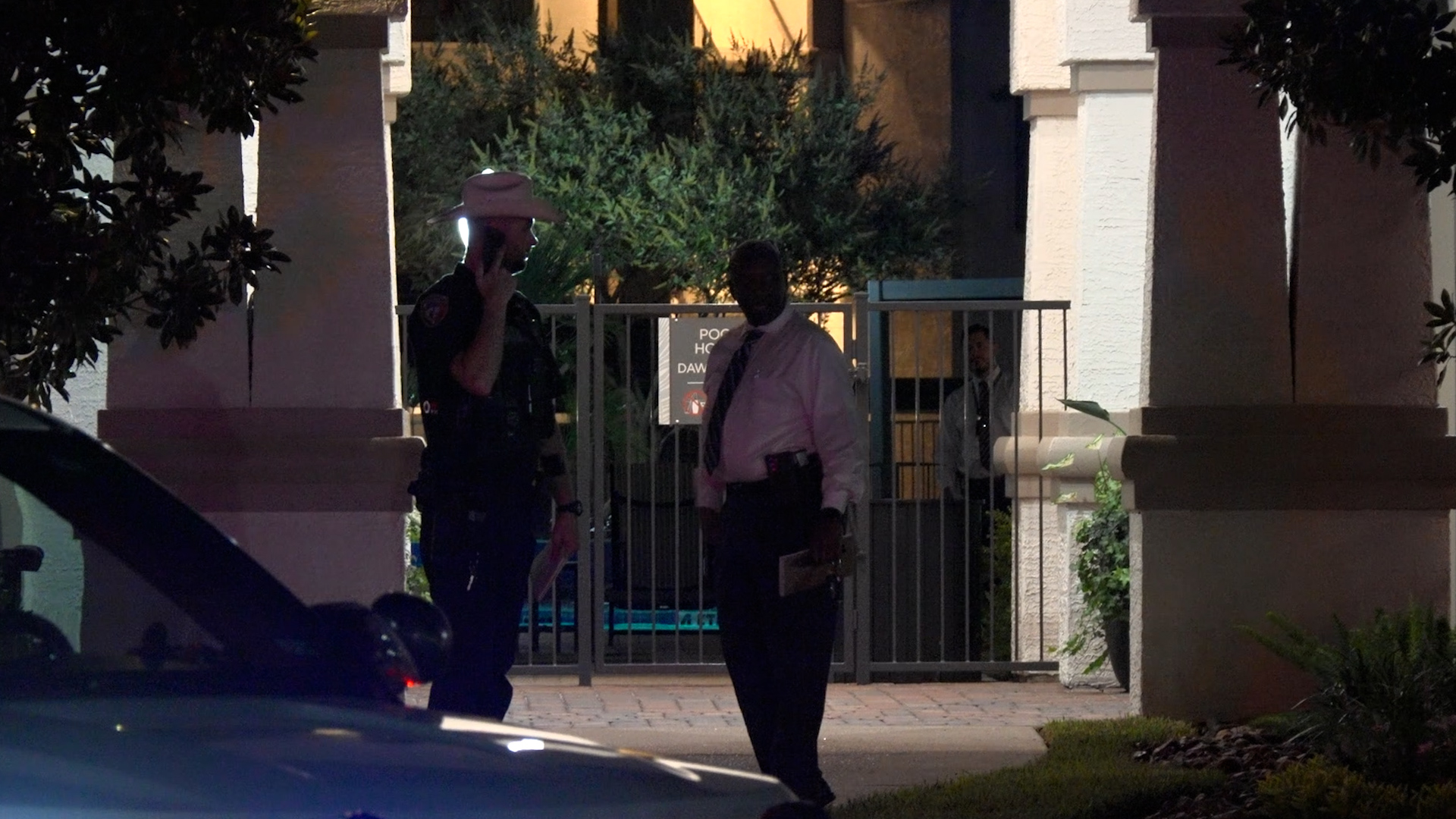 Nighttime scene of two police officers at a gated entrance, one on a cellphone, the other holding a folder near a building.