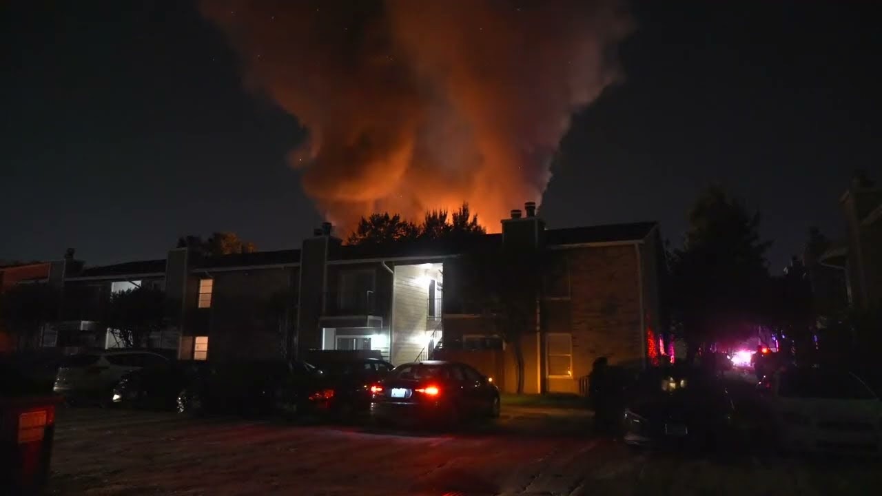 Nighttime scene of an apartment complex with a large orange fire and thick smoke rising behind trees; emergency lights glow on the right as responders arrive and cars are parked in the lot.