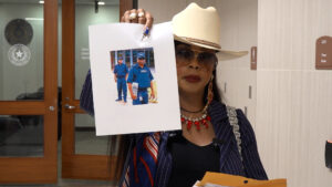 Woman in a tan hat and sunglasses holds a large printed photo in front of her, standing in a hallway with government seals visible behind her.