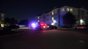 Nighttime scene in a residential parking lot with police cars and red/blue flashing lights near an apartment building.