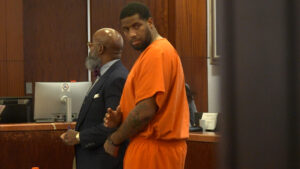 Prisoner in orange jumpsuit with tattooed arm standing beside a bearded man in a suit in a wood-paneled room.