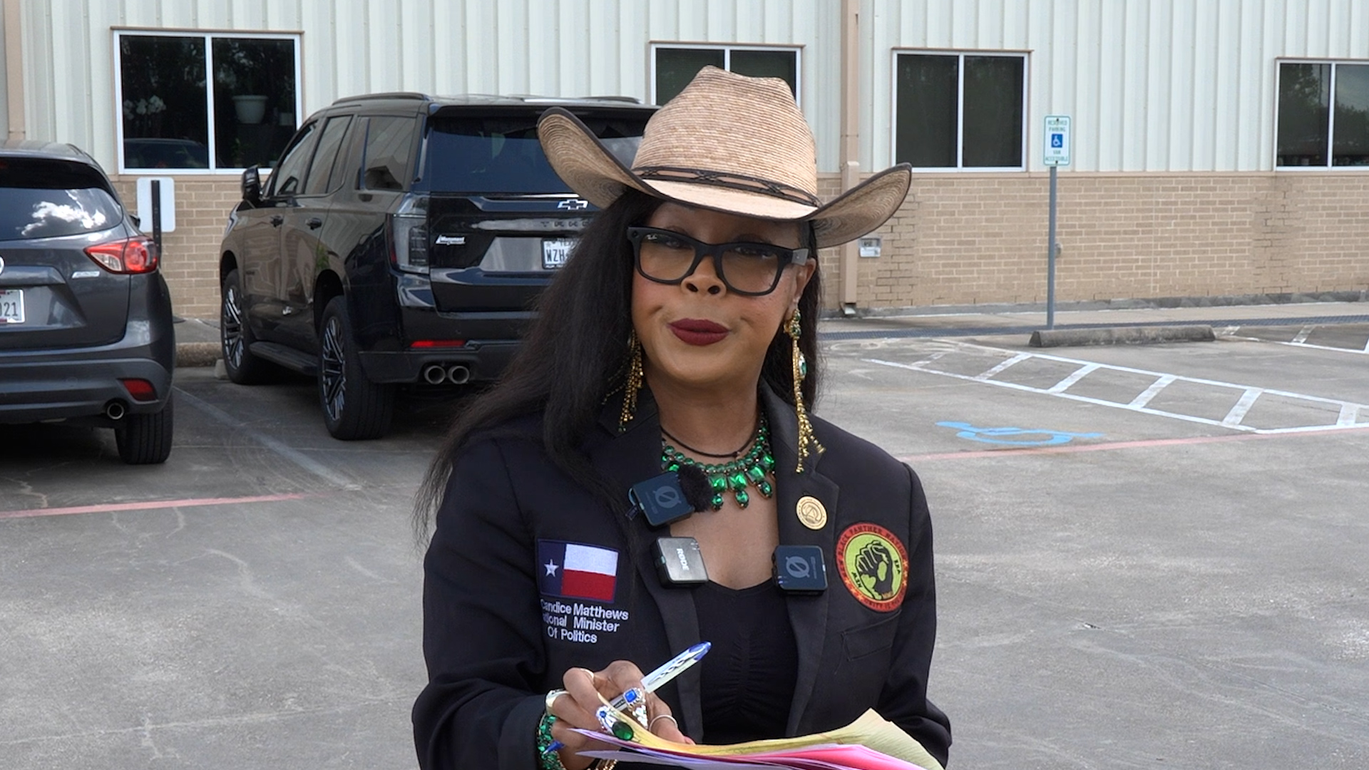 Woman wearing a tan cowboy hat, black blazer, and glasses in a parking lot, holding papers and a pen.