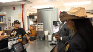 Cashier wearing a black uniform and cap speaks to customers at a fast-food counter, with a beverage dispenser and menu boards in the background.