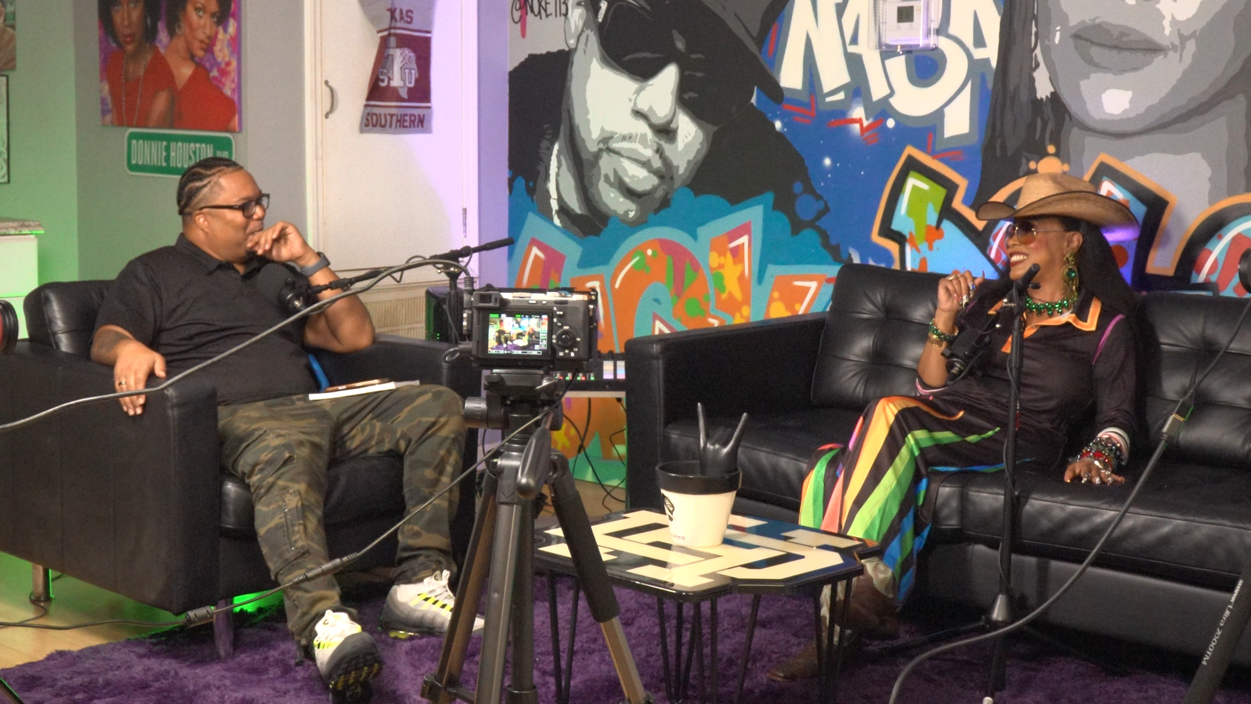 Two people sit on black couches in a colorful graffiti studio, talking into microphones during a podcast.2