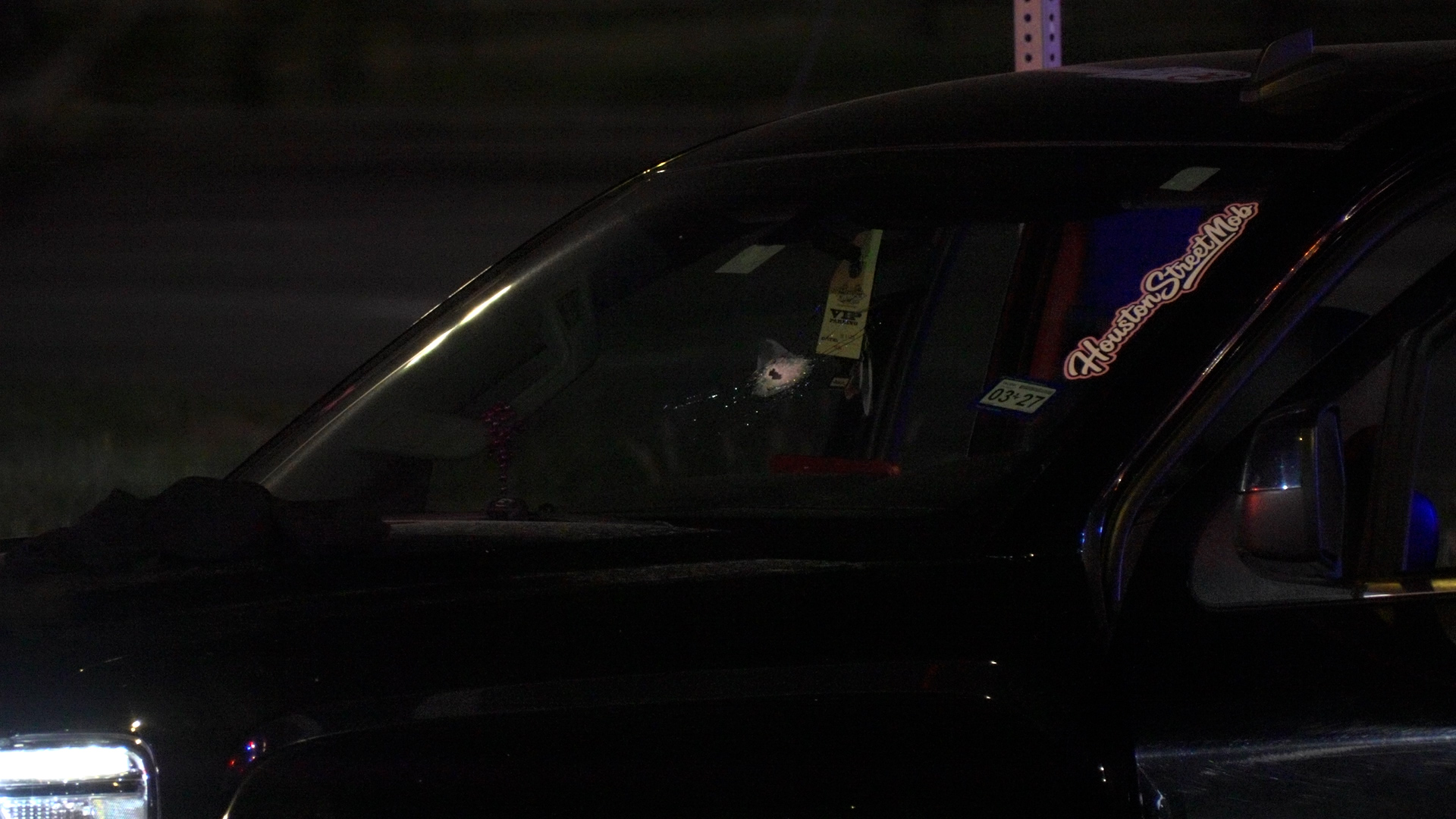 Dark car at night with a cracked windshield on the driver's side, small impact hole visible and hanging items inside the front area. stickers are on the passenger window nearby.