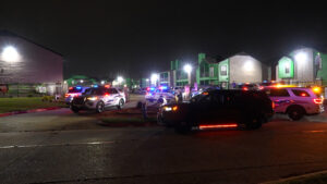 Nighttime scene with police and emergency vehicles flashing red and blue lights in a residential parking lot near green-colored apartment buildings