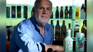 Older man in a blue shirt sits at a bar with illuminated bottles on shelves behind him.