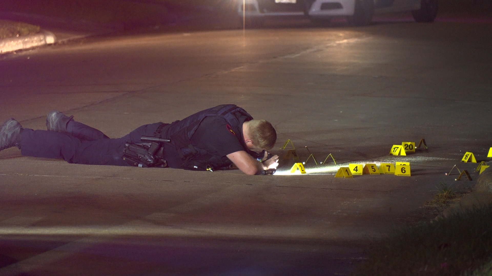 Police officer lying on a dark street at night, examining yellow evidence markers scattered on the pavement.