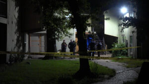 Nighttime crime scene outside a residential building with yellow caution tape and several police officers guarding the entrance at night.