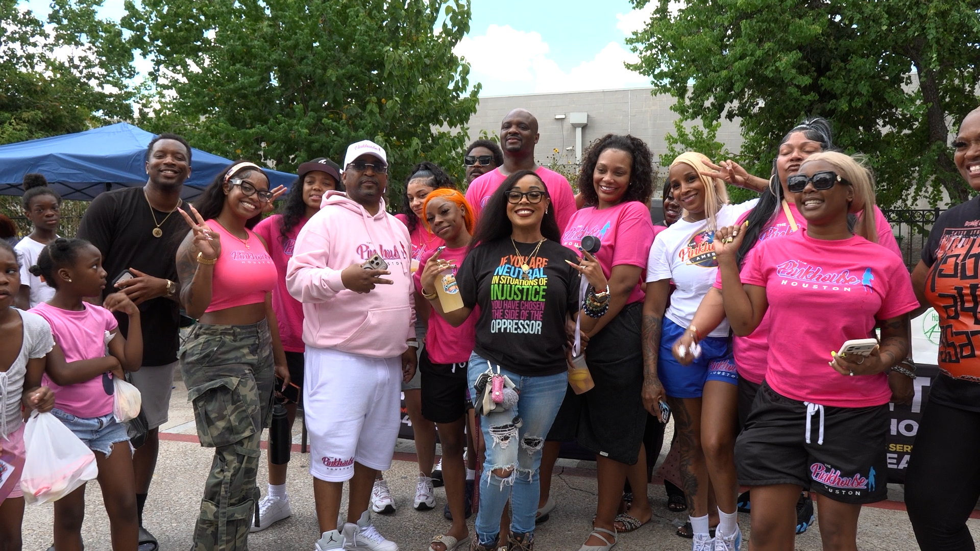 Group of people wearing pink shirts posing together at an outdoor event, smiling for the camera in a sunny setting.