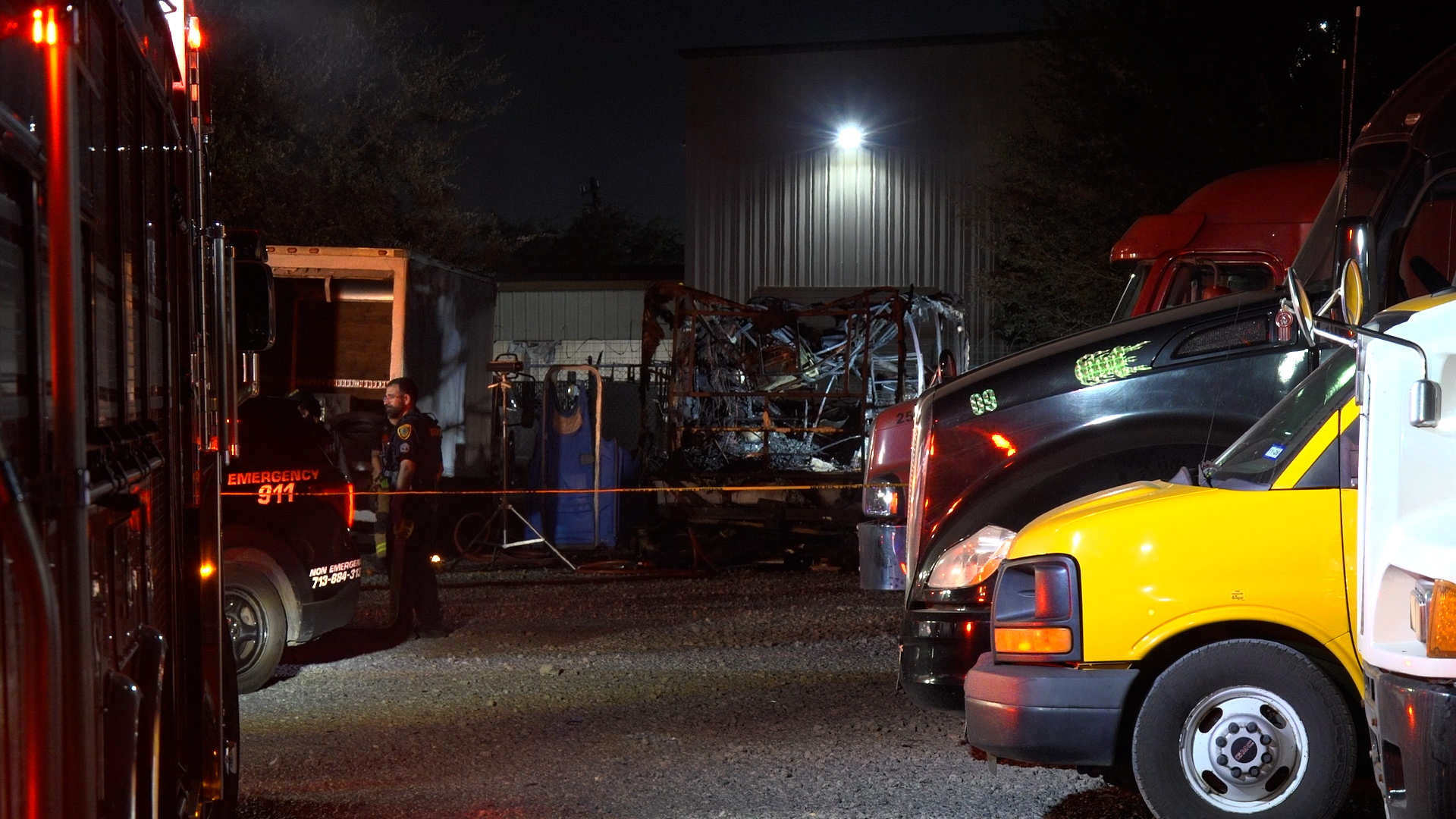 Nighttime emergency scene with fire trucks, responders, and a burned wreckage behind a fenced yard.