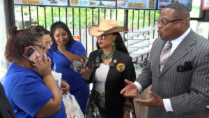 Businessman in a pinstripe suit speaks to a group of women in blue uniforms at a counter, several microphones aimed at him as they chat in a casual setting.