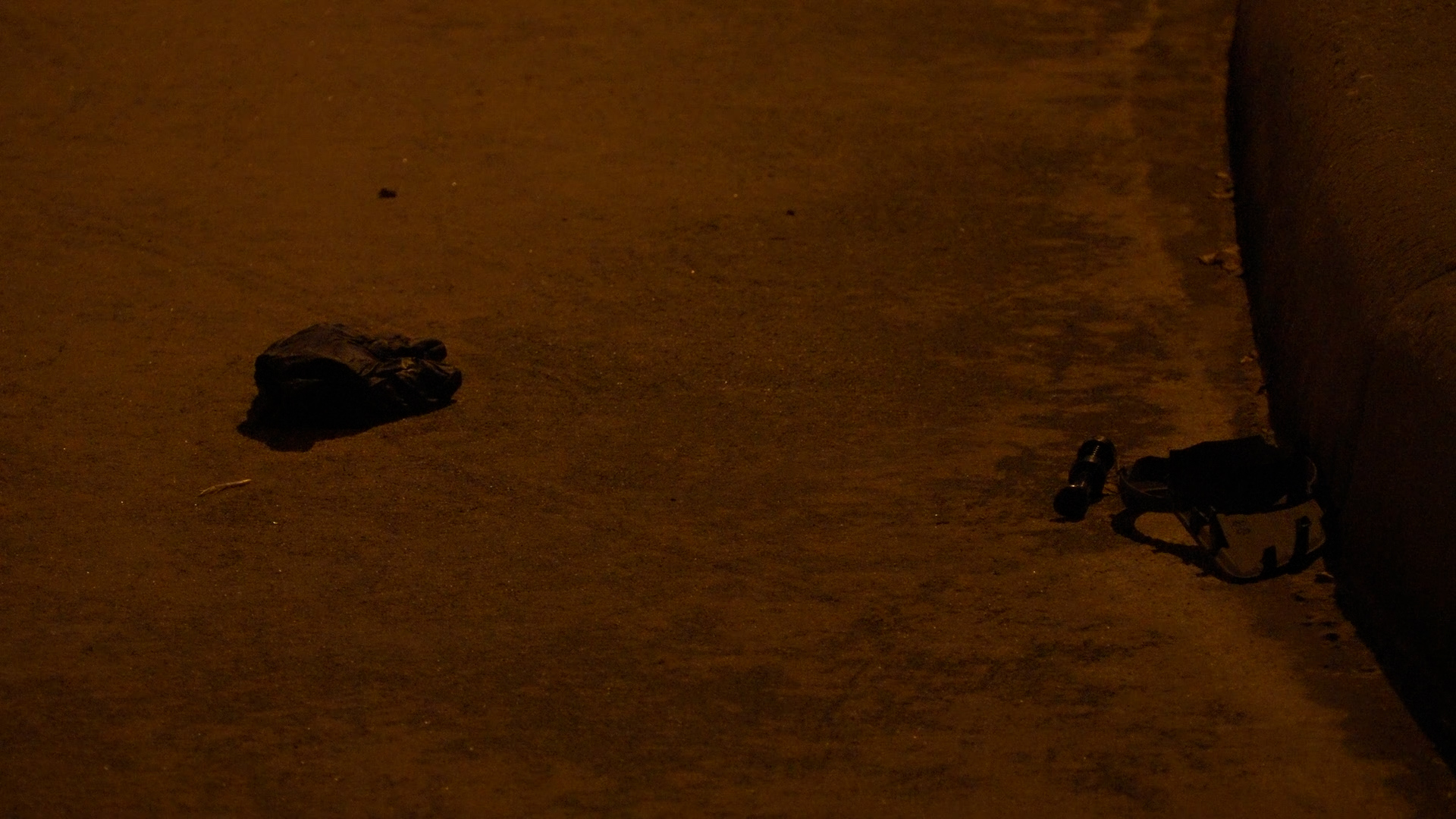 Dark street at night with a crumpled bag of trash on the ground and a helmet and bottle near a wall.