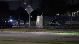 Nighttime crash scene with two damaged cars, debris on the road, and responders evaluating the incident outside a fenced area.