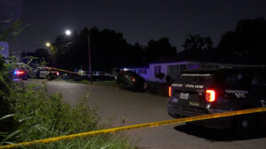 Nighttime police crime scene on a residential street: a car overturned, police SUVs with lights, and yellow caution tape cordoning the area near houses and trees.