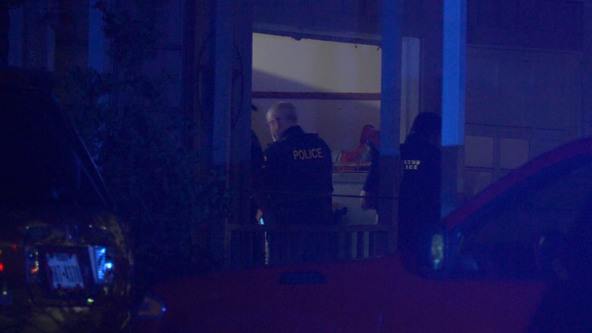 Two police officers in dark uniforms stand by an open doorway at night while blue lights illuminate the scene.