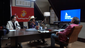 Three people sit around a large conference table in a studio podcast set, with a US Marine Corps flag on the wall and a blue screen reading 'Swimming with the Sharks' in the background, microphones and props on the table.