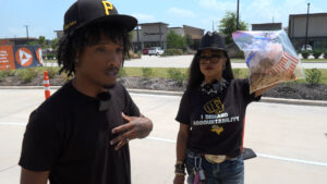 Two people stand outdoors in a parking lot; a man in a black T-shirt on the left and a woman wearing a black cowboy hat and glasses holding a large bag of chips toward the camera.