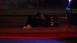 Night scene of a crashed motorcycle on the road with debris scattered and flashing emergency lights from a nearby vehicle in the background